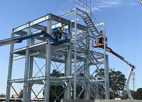 Construction workers on cherry pickers work on a metal framework of a multi-level structure. A staircase is visible, and trees are in the background under a clear blue sky.