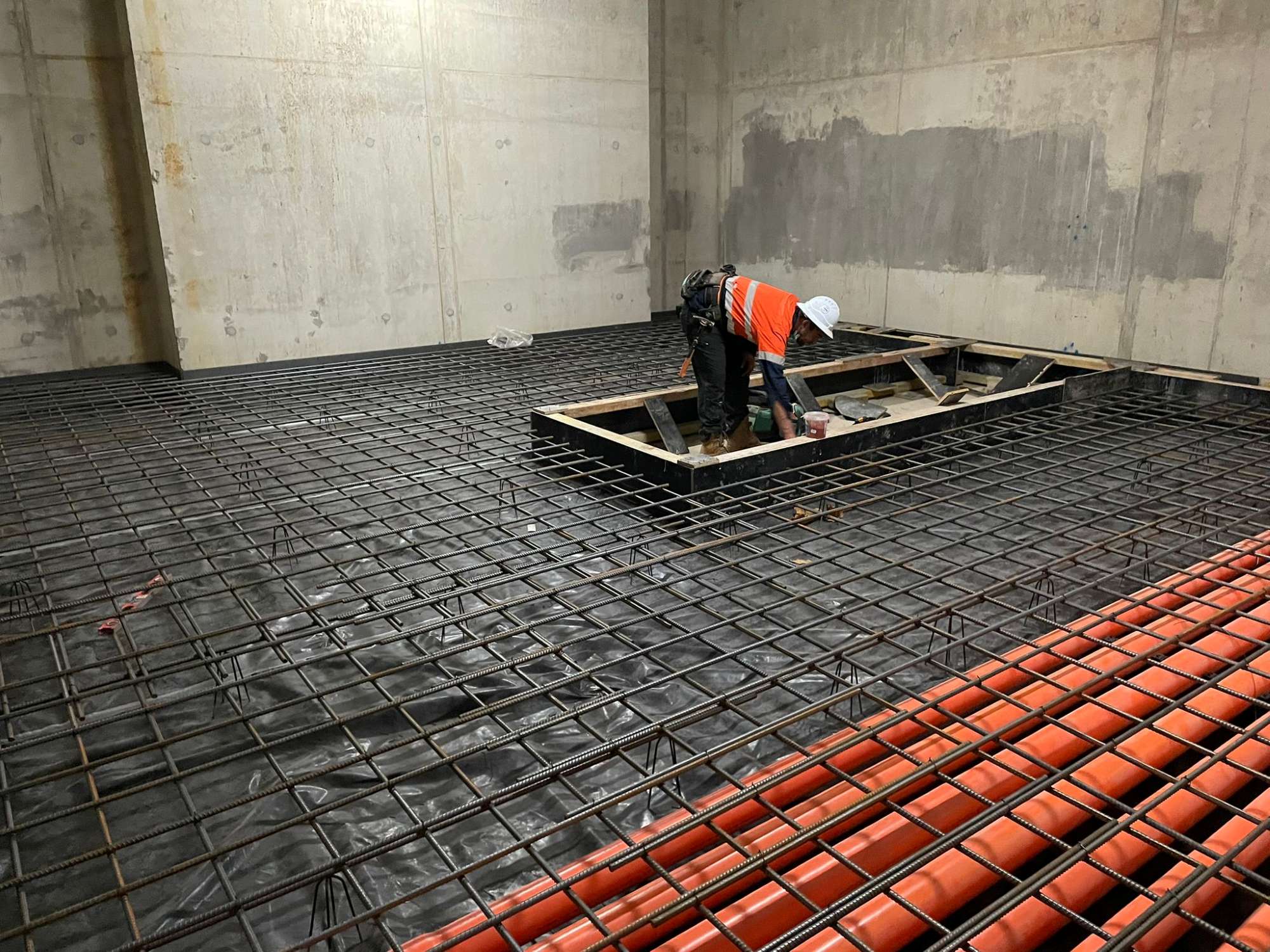 A construction worker in a white helmet and orange vest is working on a large steel rebar framework inside a concrete room. Red pipes are visible on the right side, and there's a central square wooden structure on the floor.