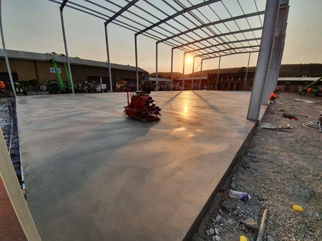 A construction site with a concrete floor being polished by a machine under a steel framework. The sun is setting in the background, casting a warm glow over the scene. Workers and equipment are visible near the structure.