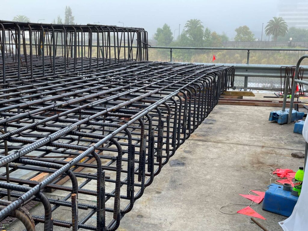 Construction site with a grid of steel reinforcement bars laid out horizontally on a concrete surface. Several orange safety cones and equipment are visible in the background, with trees and a foggy sky in the distance.