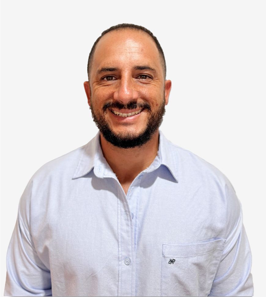 A smiling man with a short beard and short hair is wearing a light blue button-up shirt. He stands against a plain white background.
