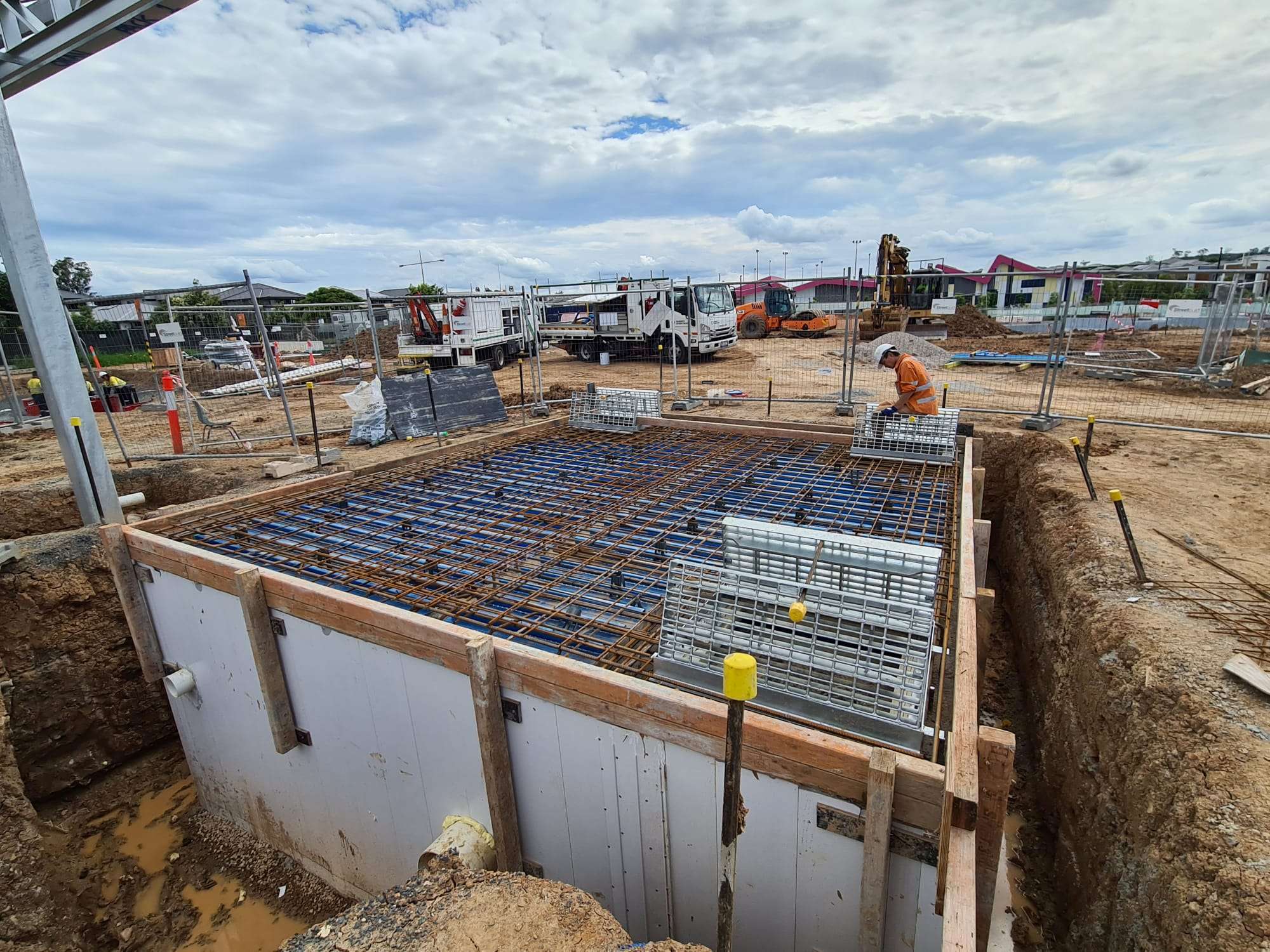 A construction site features a large foundation pit with a metal rebar grid. Surrounding equipment includes trucks, cranes, and fencing. A lone worker from Trizym Group in high-visibility clothing and helmet inspects the area under a partly cloudy sky, exemplifying promising careers in engineering projects.