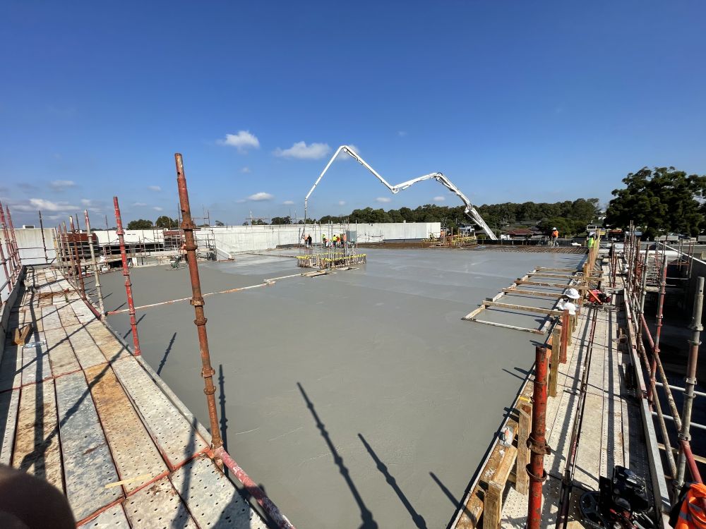 Construction site with workers on a freshly poured concrete slab. A large concrete pump is positioned over the site. Scaffolding surrounds the area, and trees are visible in the background under a clear blue sky.