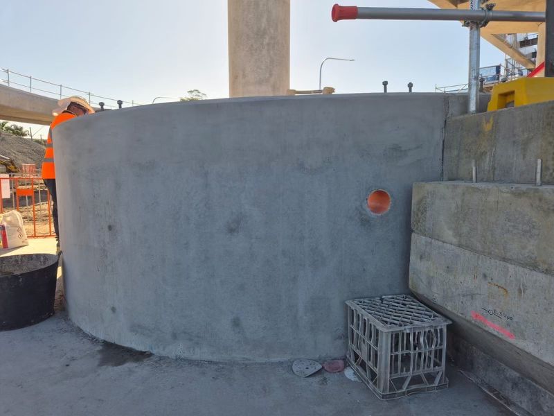 A construction site featuring a large concrete wall with a round orange pipe embedded in it. An orange-vested worker and construction materials are visible in the background. A gray crate sits on the ground beside the wall.