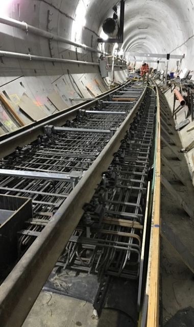 Construction of a tunnel with exposed reinforcing steel and partially completed concrete tracks. Workers and equipment are visible in the background, highlighting ongoing infrastructure development.