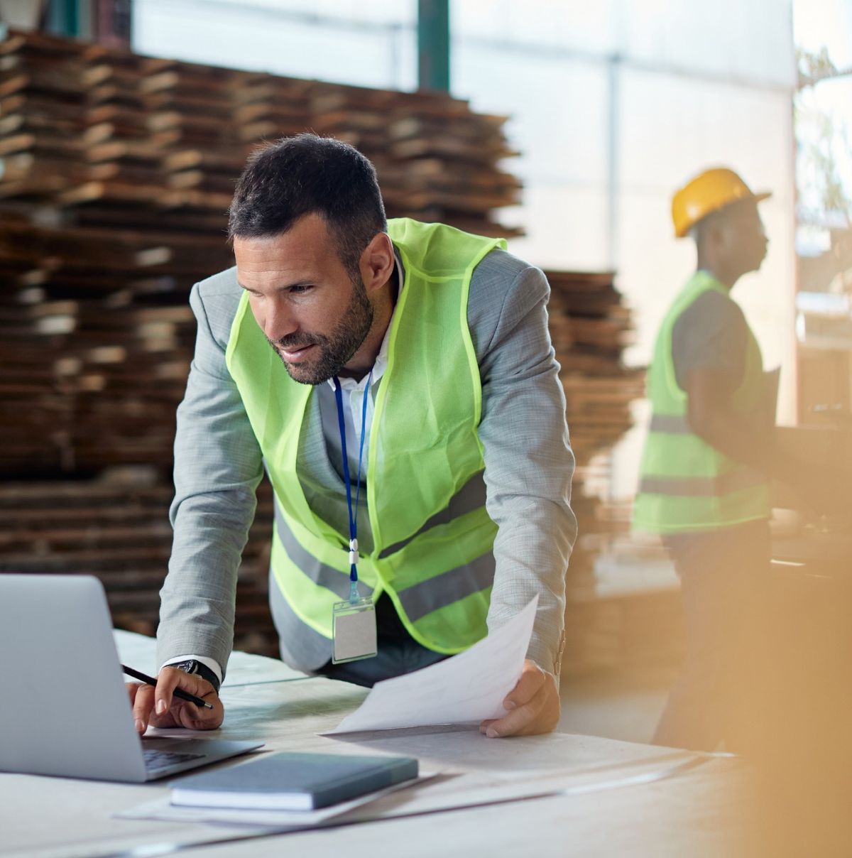 A man wearing a lime green safety vest and suit leans over a table, focused on a laptop while holding papers. In the background, another person in a safety vest and hard hat is moving through a workshop with wooden boards stacked nearby.