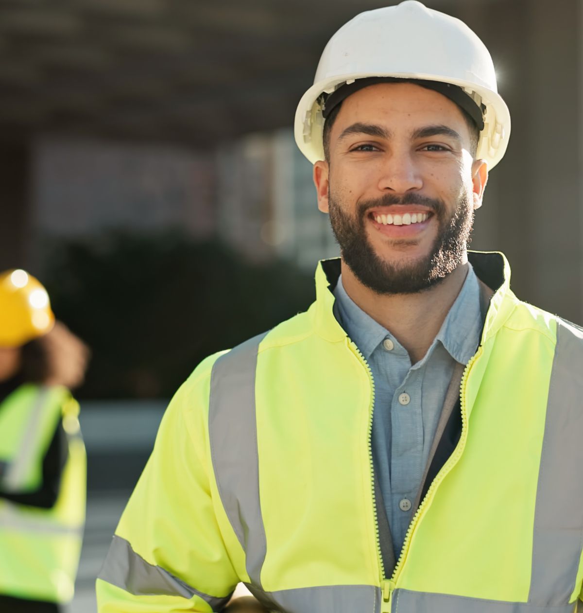 A man wearing a white hard hat and yellow reflective jacket smiles at the camera. Another worker with a hard hat is blurred in the background. They appear to be at a construction site.