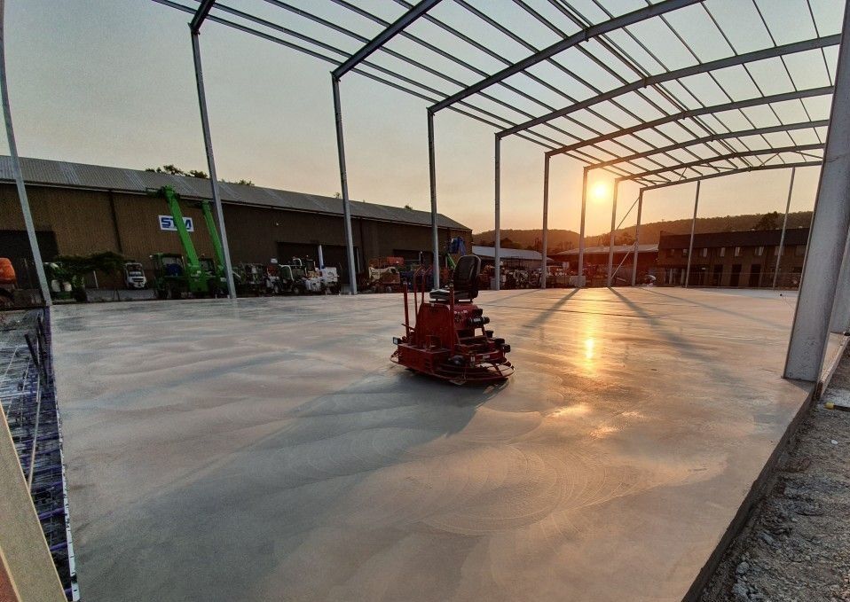 A construction site showcases a steel framework and freshly completed concrete work, with a smooth floor glistening in the warm glow of the setting sun. A small red machine rests on the floor, while buildings and equipment are visible in the distance.