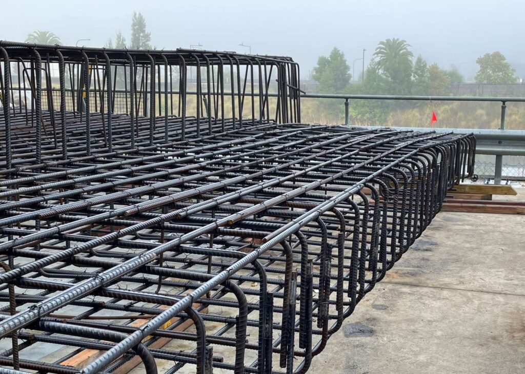 A large, intricate metal rebar structure is laid out on a concrete surface, enveloped in misty surroundings with trees in the background. This grid pattern is meticulously crafted for concrete work, likely pivotal to upcoming construction projects.