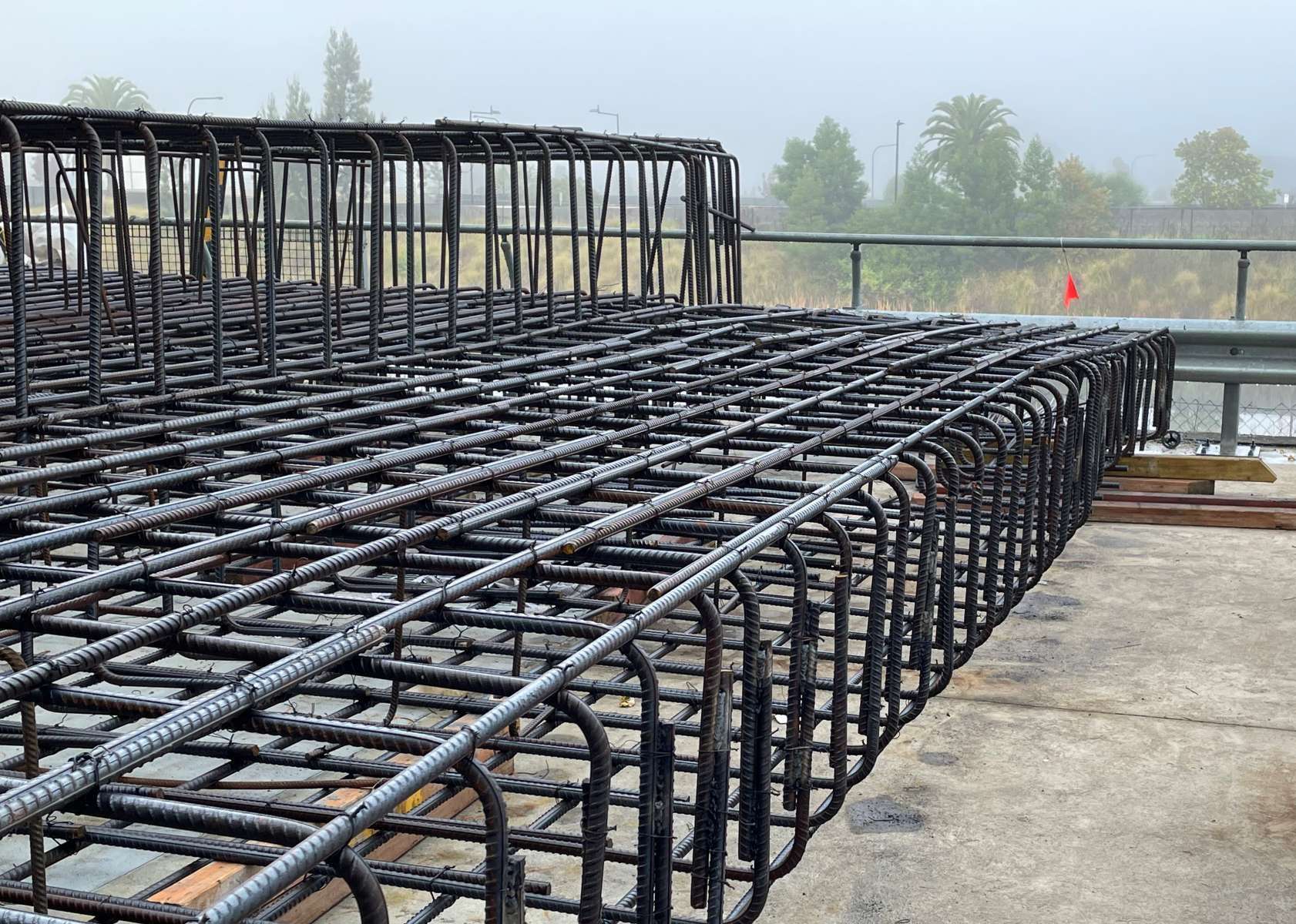 A large, intricate metal rebar structure is laid out on a concrete surface, enveloped in misty surroundings with trees in the background. This grid pattern is meticulously crafted for concrete work, likely pivotal to upcoming construction projects.